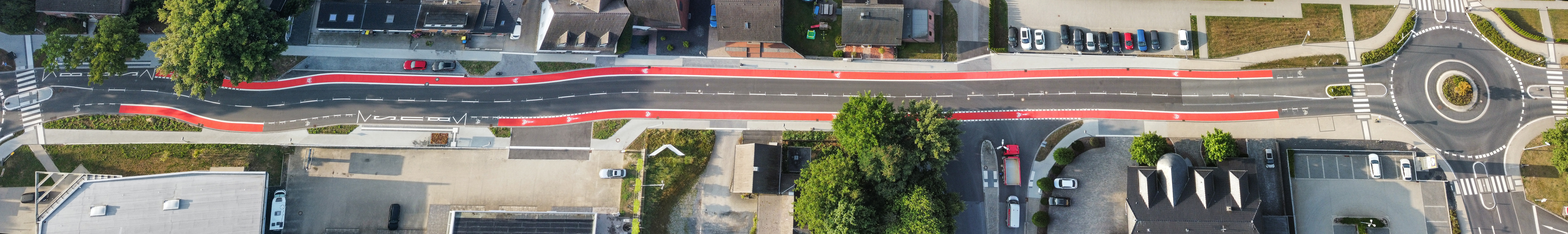 Das Bild zeigt eine zweispurige Straße aus der Vogelperspektive, die sowohl links als auch rechts durch jeweils einen Kreisverkehr begrenzt wird. Oberhalb und unterhalb der Straße befinden sich mehrere Gebäude, Fahrzeuge, Grünstreifen und Bäume. Entlang beider Straßenseiten befinden sich rot hervorgehobene Fahrradschutzstreifen. Südlich grenzt an die Straße eine Bushaltestelle.